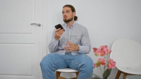 Téléchargez les photos : Jeune homme hispanique envoyant un message vocal par smartphone assis sur une chaise dans la salle d'attente - en image libre de droit