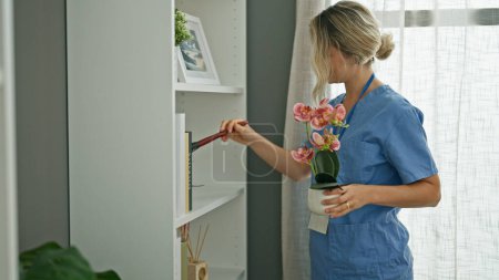 Foto de Joven mujer rubia limpiador profesional estanterías de limpieza con plumero en casa - Imagen libre de derechos