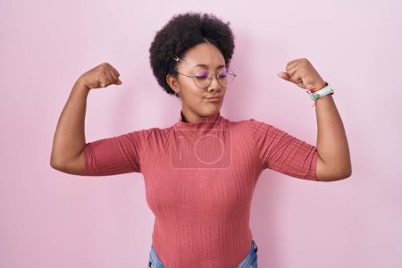 Téléchargez les photos : Belle femme africaine avec les cheveux bouclés debout sur fond rose montrant les muscles des bras souriant fier. concept de fitness. - en image libre de droit