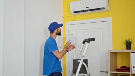 Photo for Young hispanic man technician repairing air condition machine using remote control at home - Royalty Free Image