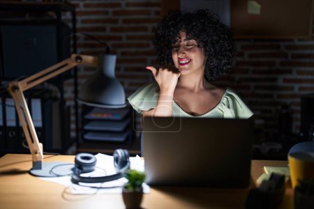 Téléchargez les photos : Jeune femme brune aux cheveux bouclés travaillant au bureau la nuit souriant avec un visage heureux regardant et pointant vers le côté avec le pouce vers le haut. - en image libre de droit