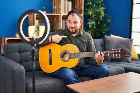 Téléchargez les photos : Plus taille hispanique homme avec barbe jouer de la guitare classique à l'enregistrement studio de musique lui-même pointant du doigt un sourire heureux et fier - en image libre de droit