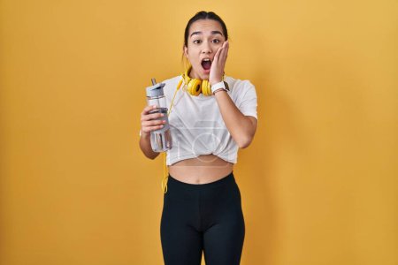 Photo for Young south asian woman wearing sportswear drinking water afraid and shocked, surprise and amazed expression with hands on face - Royalty Free Image