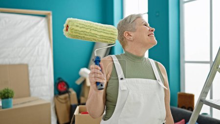 Foto de Mujer de pelo gris de mediana edad sonriendo confiado sosteniendo rodillo de pintura mirando a su alrededor en el nuevo hogar - Imagen libre de derechos