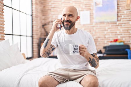 Foto de Young bald man talking on smartphone drinking coffee at bedroom - Imagen libre de derechos