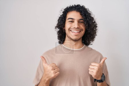 Photo for Hispanic man with curly hair standing over white background success sign doing positive gesture with hand, thumbs up smiling and happy. cheerful expression and winner gesture. - Royalty Free Image