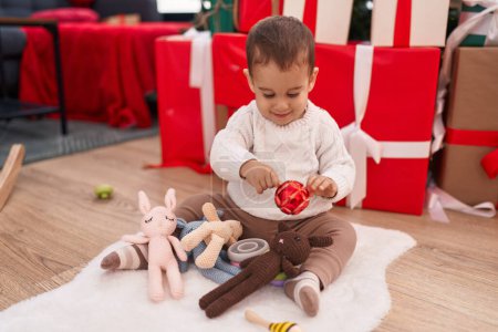 Téléchargez les photos : Adorable bambin hispanique tenant décoration de boule de Noël assis sur le sol à la maison - en image libre de droit