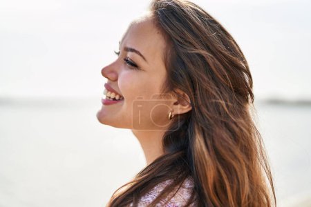 Téléchargez les photos : Jeune belle hispanique femme touriste souriant confiant regardant sur le côté à la plage - en image libre de droit