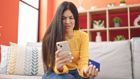 Téléchargez les photos : Jeune belle hispanique femme shopping avec smartphone et carte de crédit assis sur le canapé à la maison - en image libre de droit