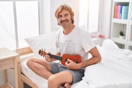 Téléchargez les photos : Jeune homme blond jouant ukulele assis sur le lit dans la chambre - en image libre de droit