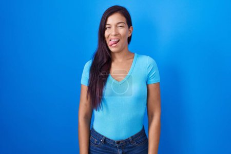 Foto de Mujer hispana joven de pie sobre fondo azul sacando la lengua feliz con expresión divertida. concepto de emoción. - Imagen libre de derechos