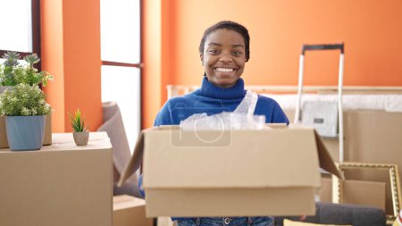 Téléchargez les photos : Afro-américaine femme souriant confiant tenant paquet à nouveau à la maison - en image libre de droit