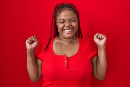 Foto de Mujer afroamericana con el pelo trenzado de pie sobre fondo rojo emocionada por el éxito con los brazos levantados y los ojos cerrados celebrando la victoria sonriendo. concepto ganador. - Imagen libre de derechos