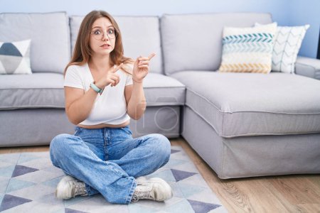 Photo for Young caucasian woman sitting on the floor at the living room pointing aside worried and nervous with both hands, concerned and surprised expression - Royalty Free Image