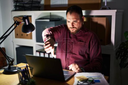 Photo for Plus size hispanic man with beard working at the office at night looking unhappy and angry showing rejection and negative with thumbs down gesture. bad expression. - Royalty Free Image