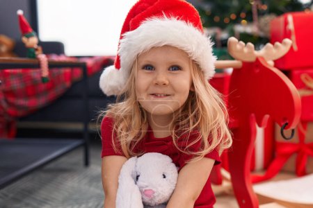 Téléchargez les photos : Adorable fille blonde souriante confiante assise sur le sol près de l'arbre de Noël à la maison - en image libre de droit