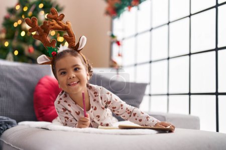 Foto de Adorable chica hispana sonriendo confiada acostada en el sofá por el árbol de Navidad en casa - Imagen libre de derechos