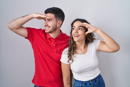 Photo for Young hispanic couple standing over isolated background very happy and smiling looking far away with hand over head. searching concept. - Royalty Free Image