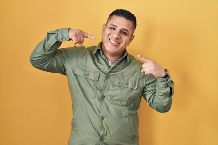 Téléchargez les photos : Jeune homme hispanique debout sur fond jaune souriant gai montrant et pointant avec les doigts dents et la bouche. concept de santé dentaire. - en image libre de droit