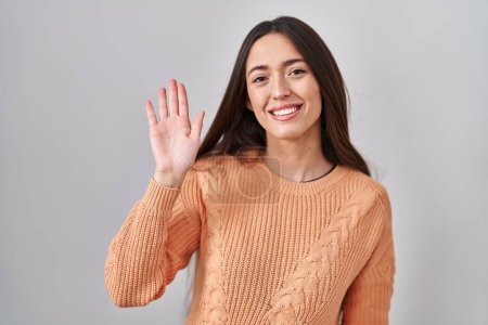 Téléchargez les photos : Jeune femme brune debout sur fond blanc renonçant à dire bonjour heureux et souriant, geste de bienvenue amical - en image libre de droit