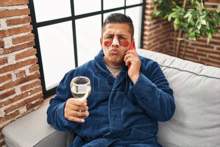 Photo for Hispanic young man wearing bathrobe and eye bags patches drinking wine speaking on the phone looking at the camera blowing a kiss being lovely and sexy. love expression. - Royalty Free Image