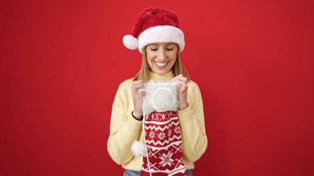 Téléchargez les photos : Jeune femme blonde portant un chapeau de Noël regardant à l'intérieur de la chaussette sur fond rouge isolé - en image libre de droit