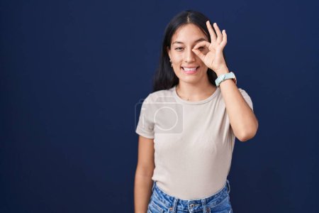 Foto de Mujer hispana joven de pie sobre fondo azul haciendo buen gesto con la mano sonriente, los ojos mirando a través de los dedos con cara feliz. - Imagen libre de derechos