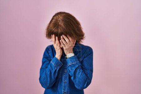 Foto de Mujer de mediana edad de pie sobre fondo rosa con expresión triste cubriendo la cara con las manos mientras llora. concepto de depresión. - Imagen libre de derechos