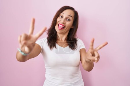 Téléchargez les photos : Femme brune du moyen âge debout sur fond rose souriant avec la langue montrant les doigts des deux mains faisant signe de victoire. numéro deux. - en image libre de droit