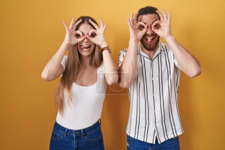 Photo for Young couple standing over yellow background doing ok gesture like binoculars sticking tongue out, eyes looking through fingers. crazy expression. - Royalty Free Image