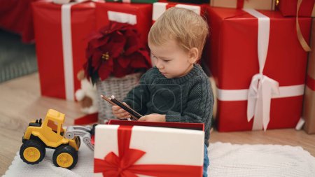 Téléchargez les photos : Adorable tout-petit blond déballer cadeau de Noël assis sur le sol à la maison - en image libre de droit