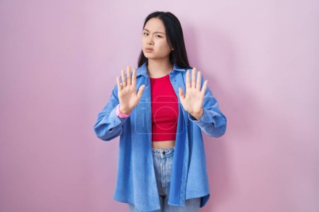Foto de Mujer asiática joven de pie sobre fondo rosa alejando las palmas de las manos mostrando rechazo y negación con miedo y expresión repugnante. parada y prohibido. - Imagen libre de derechos