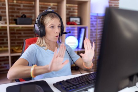 Photo for Young caucasian woman playing video games wearing headphones moving away hands palms showing refusal and denial with afraid and disgusting expression. stop and forbidden. - Royalty Free Image