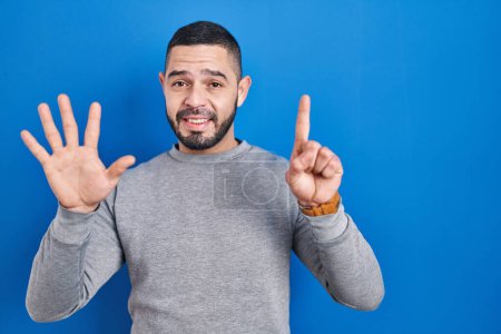 Foto de Hombre hispano de pie sobre fondo azul mostrando y señalando hacia arriba con los dedos número seis mientras sonríe confiado y feliz. - Imagen libre de derechos