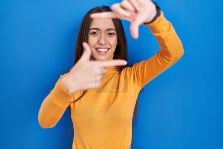 Téléchargez les photos : Young brunette woman standing over blue background smiling making frame with hands and fingers with happy face. creativity and photography concept. - en image libre de droit