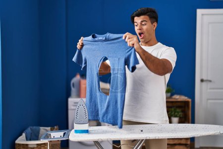 Photo for Hispanic man ironing holding burned iron shirt at laundry room celebrating crazy and amazed for success with open eyes screaming excited. - Royalty Free Image