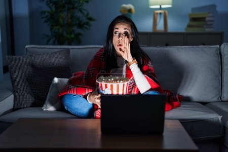 Téléchargez les photos : Femme hispanique manger du pop-corn regarder un film sur le canapé main sur la bouche dire rumeur secrète, chuchoter conversation malicieuse - en image libre de droit
