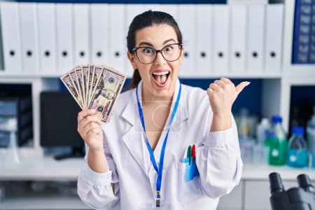Téléchargez les photos : Jeune femme brune travaillant au laboratoire scientifique tenant de l'argent pointant le pouce vers le côté souriant heureux avec la bouche ouverte - en image libre de droit