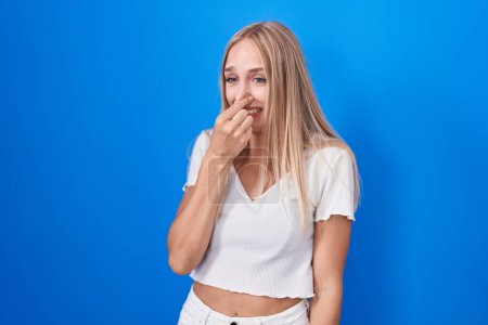 Foto de Young caucasian woman standing over blue background smelling something stinky and disgusting, intolerable smell, holding breath with fingers on nose. bad smell - Imagen libre de derechos