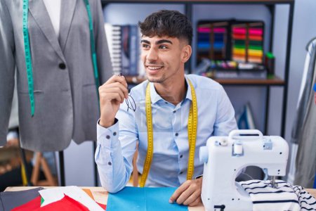 Téléchargez les photos : Jeune homme hispanique tailleur souriant confiant assis sur la table au magasin de tailleur - en image libre de droit