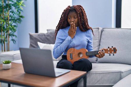 Téléchargez les photos : Femme africaine jouant ukulélé à la maison faire appel vidéo couvrant la bouche avec la main, choqué et peur de l'erreur. expression surprise - en image libre de droit