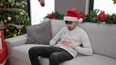 Téléchargez les photos : Jeune homme hispanique souffrant de maux d'estomac assis près de l'arbre de Noël à la maison - en image libre de droit