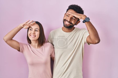 Foto de Jóvenes pareja hispana juntos sobre fondo rosa muy feliz y sonriente mirando lejos con la mano sobre la cabeza. concepto de búsqueda. - Imagen libre de derechos