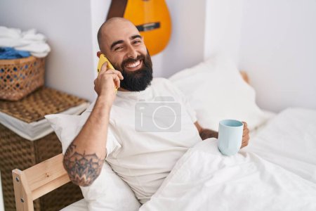 Foto de Young bald man talking on smartphone drinking coffee at bedroom - Imagen libre de derechos