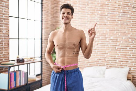 Photo for Young hispanic man using tape measure measuring waist smiling happy pointing with hand and finger to the side - Royalty Free Image