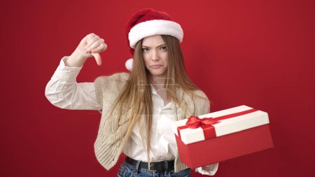Téléchargez les photos : Jeune femme blonde portant chapeau de Noël tenant cadeau regardant bouleversé sur fond rouge isolé - en image libre de droit