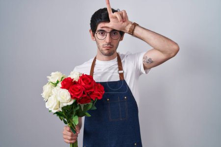 Téléchargez les photos : Jeune homme hispanique tenant un bouquet de roses blanches et rouges se moquant des gens avec les doigts sur le front faisant geste de perdant se moquant et insultant. - en image libre de droit