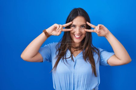 Téléchargez les photos : Young brunette woman standing over blue background doing peace symbol with fingers over face, smiling cheerful showing victory - en image libre de droit