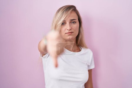 Photo for Young blonde woman standing over pink background looking unhappy and angry showing rejection and negative with thumbs down gesture. bad expression. - Royalty Free Image