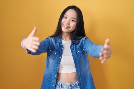 Foto de Joven mujer asiática de pie sobre fondo amarillo mirando a la cámara sonriendo con los brazos abiertos para abrazar. expresión alegre abrazando la felicidad. - Imagen libre de derechos